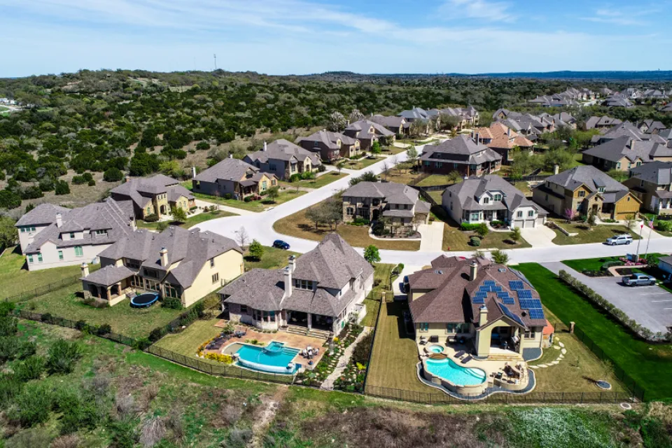 A subdivision of large suburban homes, some with bright blue pools, sits atop a low hill in Hays County, Texas.